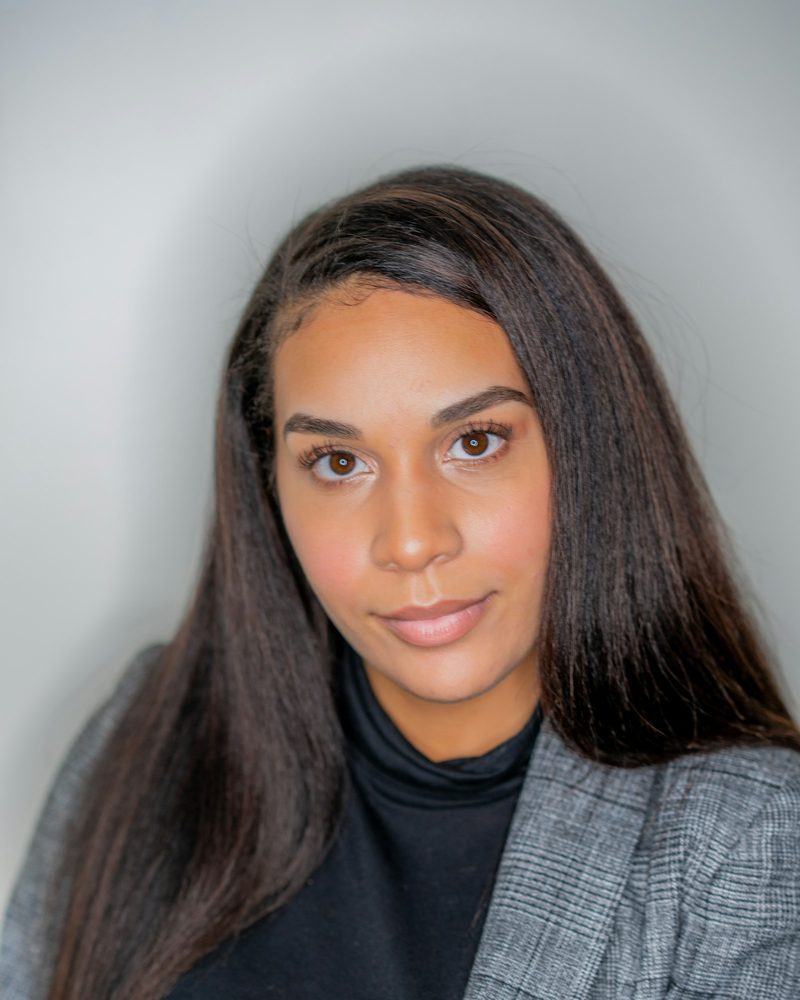 Portrait von Mara mit langen dunklen Haaren, schwarzem Rollkragen und grauem Blazer vor hellgrauem Hintergrund